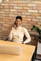 1 jovem africano americano o negócio mulher usando Móvel telefone enquanto sentado de a tijolo parede dentro a industrial estilo escritório foto