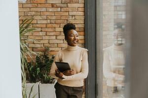 1 jovem africano americano o negócio mulher com digital tábua em pé de a tijolo parede dentro a industrial estilo escritório foto