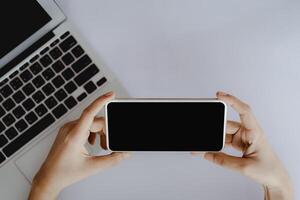 topo Visão do mão segurando Smartphone com em branco Preto tela e computador computador portátil em branco escritório escrivaninha foto