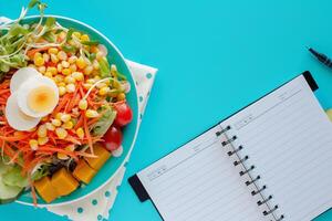 fresco salada vegetal com fervido frango ovo, em branco espiral caderno e uma caneta em azul fundo foto