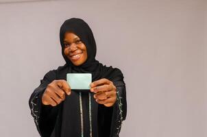 lindo muçulmano mulher segurando crédito cartão e sorridente às Câmera isolado em cinzento foto