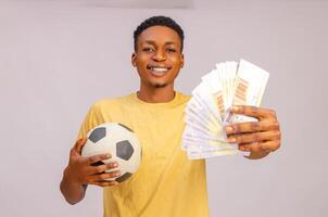 esporte apostas. retrato do animado Preto cara com futebol bola e dinheiro em pé sobre bege fundo. foto