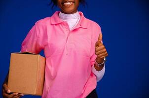 lindo africano fêmea correio trabalhador segurando uma caixa, faz uma polegares acima gesto foto