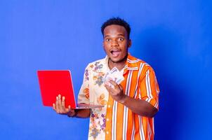 foto do uma feliz jovem afro americano bonito homem posando isolado sobre azul parede fundo usando computador portátil computador segurando dinheiro.