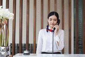 jovem ásia mulher recepcionista atrás a hotel contador falando em a telefone, fêmea recepcionista trabalhando dentro a hotel foto