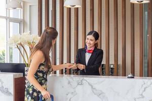 jovem ásia mulher recepcionista às a hotel contador falando com borrado fêmea Viajantes check-in às a hotel, fêmea recepcionista trabalhando dentro a hotel foto