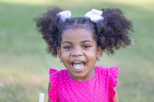 feliz pré-escolar africano menina degola dele língua Fora com doce, criança menina jogando ao ar livre dentro a parque foto