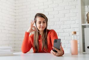 jovem mulher caucasiana usando fones de ouvido, conversando no celular, mostrando sinal de ok foto