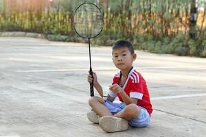 ásia Garoto sentado de pernas cruzadas em a cimento chão mão segurando uma branco peteca. a de outros mão detém uma badminton raquete. ela descansado enquanto jogando badminton lado de fora em dela dia desligado. foto