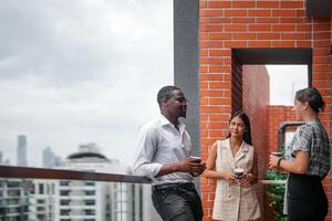 equipe do o negócio pessoas inteligente homem e mulheres ficar de pé às ao ar livre terraço construção e conversa juntos com café da manhã Comida e café em a mão dentro Boa sentindo-me com cidade espaço prédio. o negócio manhã. foto