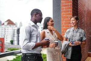 equipe do o negócio pessoas inteligente homem e mulheres ficar de pé às ao ar livre terraço construção e conversa juntos com café da manhã Comida e café em a mão dentro Boa sentindo-me com cidade espaço prédio. o negócio manhã. foto