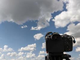 Preto profissional digital Câmera em uma tripé aguçado às azul céu com branco nuvens foto