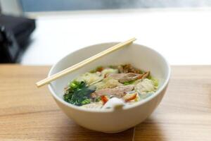 carne de porco dumplings Wonton sopa com bok choy e churrasco vermelho carne de porco servir com pauzinhos em uma tigela em de madeira mesa e luz solar fundo foto