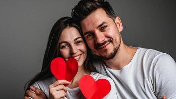 ai gerado fofa jovem casal é segurando vermelho papel corações, olhando às Câmera e sorridente enquanto em pé contra cinzento fundo foto