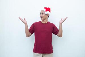 jovem ásia homem vestindo uma santa claus chapéu sorridente, choque e apontando para dela lado isolado de branco fundo para visual comunicação foto