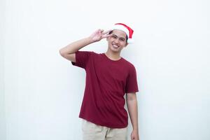 jovem ásia homem vestindo uma santa claus chapéu fazer uma Paz placa com 2 dedos isolado de uma branco fundo para visual comunicação foto