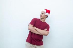 jovem ásia homem vestindo uma santa claus chapéu cruzando dele braços enquanto sorridente, choque e rindo com uma branco fundo para visual comunicação foto