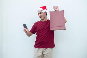 jovem ásia homem vestindo uma santa claus chapéu segurando uma Smartphone e uma compras saco com expressões do sorriso, choque, e surpresa, isolado contra uma branco fundo para visual comunicação foto