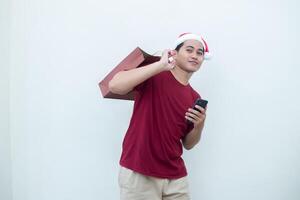 jovem ásia homem vestindo uma santa claus chapéu segurando uma Smartphone e uma compras saco com expressões do sorriso, choque, e surpresa, isolado contra uma branco fundo para visual comunicação foto