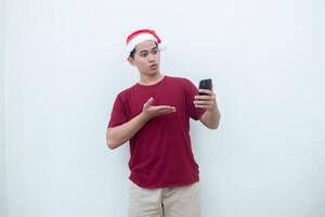 jovem ásia homem vestindo uma santa claus chapéu segurando uma Smartphone e expressando uma sorriso, surpresa, e apontando para a lado, isolado de uma branco fundo para visual comunicação foto