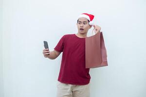 jovem ásia homem vestindo uma santa claus chapéu segurando uma Smartphone e uma compras saco com expressões do sorriso, choque, e surpresa, isolado contra uma branco fundo para visual comunicação foto