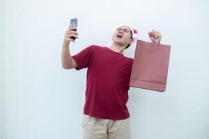 jovem ásia homem vestindo uma santa claus chapéu segurando uma Smartphone e uma compras saco com expressões do sorriso, choque, e surpresa, isolado contra uma branco fundo para visual comunicação foto