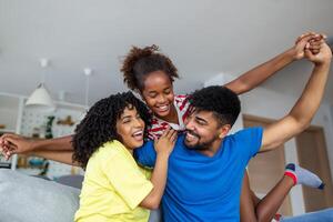 feliz africano americano Papai e mãe pegando carona animado orgulhoso filha criança, jogando vôo Super heroi, alcançando braço avançar. alegre menina equitação pais costas jogando ativo jogos com família às casa foto