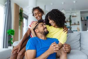 atraente feliz família abraçando atraente mãe bonito pai e adorável pequeno filha sentado juntos em sofá dentro vivo quarto às casa sorridente olhando às Câmera foto