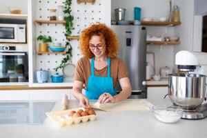 feliz atraente jovem adulto mulher dona de casa padeiro vestem avental segurando PIN rolando massa em cozinha mesa cozimento pastelaria conceito cozinhando bolo bolacha fazendo padaria fazer caseiro pizza às casa foto