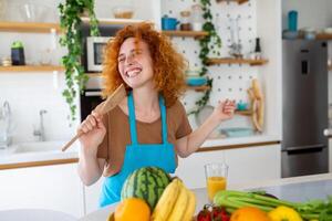 engraçado lindo mulher cantando para dentro espátula, cozinhando dentro moderno cozinha, segurando espátula Como microfone, dançando, ouvindo para música, brincalhão menina tendo Diversão com utensílios de cozinha, preparando Comida. foto