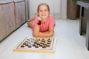 sorridente menina deitado em a chão às casa Próximo para jogo de damas pronto para jogar foto