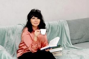 Senior mulher sentado em a sofá bebendo café e lendo uma livro foto