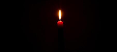 uma solteiro queimando vela chama ou luz brilhando em vermelho vela em Preto ou Sombrio fundo em mesa dentro Igreja para Natal, velório ou memorial serviço com cópia de espaço foto