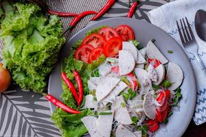 picante vidro macarrão salada com vietnamita linguiça servido dentro uma cinzento prato colocar em uma de madeira mesa e vários legumes. foto