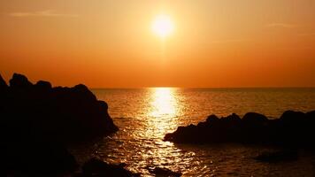a Sol é configuração em a mar com pedras dentro a primeiro plano, Preto silhuetas em a rochas, luz refletindo fora a mar. foto