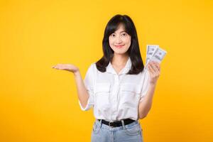 jovem feliz mulher do ásia etnia vestem branco camisa e jeans brim segurando dinheiro dinheiro dentro dólar e apontando mão para livre cópia de espaço contra amarelo fundo. investimento e financeiro conceito. foto