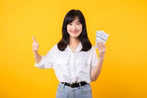 jovem feliz mulher do ásia etnia vestem branco camisa e jeans brim segurando dinheiro dinheiro dentro dólar e mostrando polegar acima gesto contra amarelo fundo. Boa investimento e financeiro conceito. foto