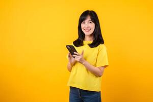 experiência a felicidade e conexão do tecnologia com alegre retrato. jovem ásia mulher vestindo amarelo camiseta e jeans jeans sorrisos usando Smartphone. conceito do tecnologia e Smartphone aplicativo. foto