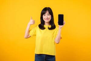 experiência positividade com retrato jovem ásia mulher vestindo uma amarelo camiseta e jeans jeans vitrines feliz sorriso, polegares acima gesto, e Smartphone tela. recomendado formulários conceito. foto