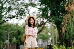 retrato do ásia jovem mulher viajante com tecelagem chapéu e cesta feliz sorrir em verde público parque natureza fundo. viagem viagem estilo de vida, mundo viagem explorador ou Ásia verão turismo conceito. foto