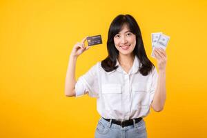 feliz bem sucedido confiante jovem ásia mulher feliz sorrir vestindo branco camisa e jeans brim segurando dinheiro dinheiro e crédito cartão em pé sobre amarelo fundo. milionário negócios, compras conceito. foto