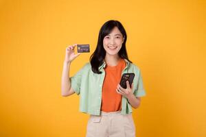 jovem ásia mulher dentro dela 30s, vestido dentro laranja camisa e verde saltador, lojas conectados usando Smartphone com crédito cartão em amarelo estúdio fundo. Móvel conceito. foto