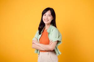 confiança e bem estar com uma jovem ásia mulher 30s vestindo laranja camisa. dela braço Cruz gesto em peito contra uma vibrante amarelo fundo tintas uma retrato do autoconfiança e interior paz. foto