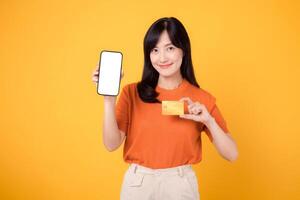 alegre ásia mulher dentro dela 30s, exibindo em branco Smartphone tela e crédito cartão em vibrante amarelo fundo. rápido conectados Forma de pagamento compras. foto