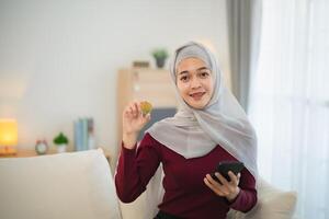 muçulmano islamismo ásia mulher vestindo hijab sorridente segurando ouro bitcoin criptografia moeda blockchain sentado em sofá dentro vivo quarto às lar. criptografia moeda blockchain salvando investimento riqueza conceito. foto