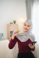 muçulmano islamismo ásia mulher vestindo hijab sorridente segurando ouro bitcoin criptografia moeda blockchain sentado em sofá dentro vivo quarto às lar. criptografia moeda blockchain salvando investimento riqueza conceito. foto