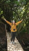 mulher vestido dentro uma amarelo Jaqueta e Preto calça, carregando uma vermelho mochila, em pé em uma de madeira ponte, com dela braços abrir, dentro a meio do uma floresta durante uma ensolarado dia foto