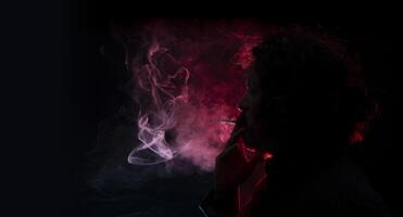 homem com uma cigarro dentro dele mão, sopro fumaça Fora do dele boca visto dentro perfil dentro silhueta com vermelho luz iluminador dele perfil contra Preto fundo foto