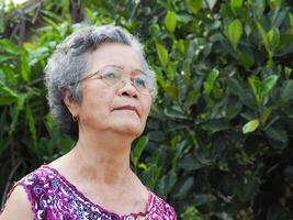 uma Senior ásia mulher com curto branco cabelo sorridente e olhando acima enquanto em pé dentro uma jardim foto