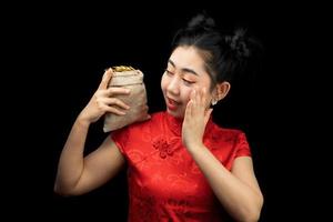 retrato asiático jovem, mulher vestido vermelho tradicional cheongsam segurando uma moeda de ouro em um saco no fundo preto foto